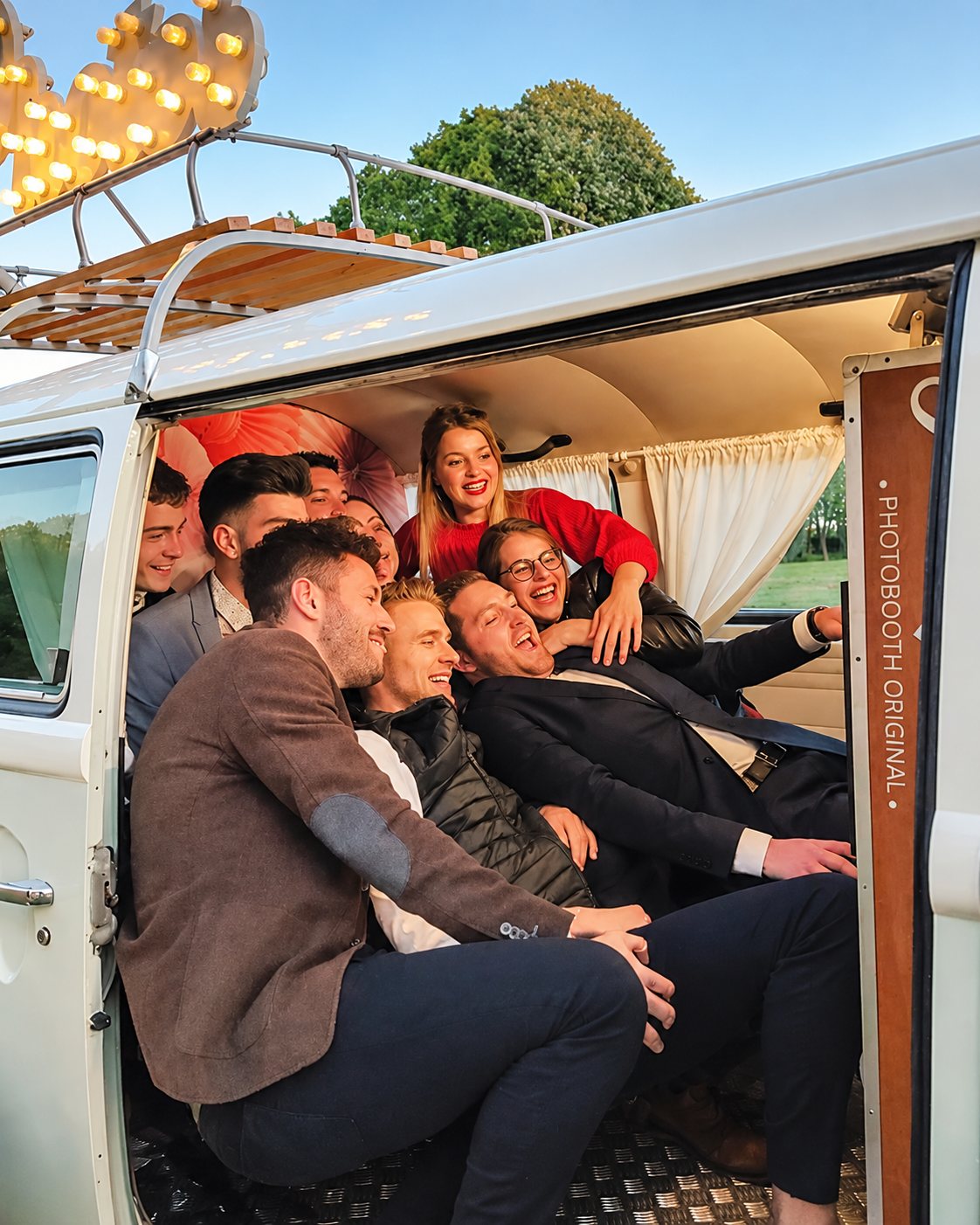 Un groupe de jeunes gens rit en se serrant à l'intérieur d'un combi Volkswagen blanc aménagé en photobooth, avec des lettres lumineuses sur le toit.