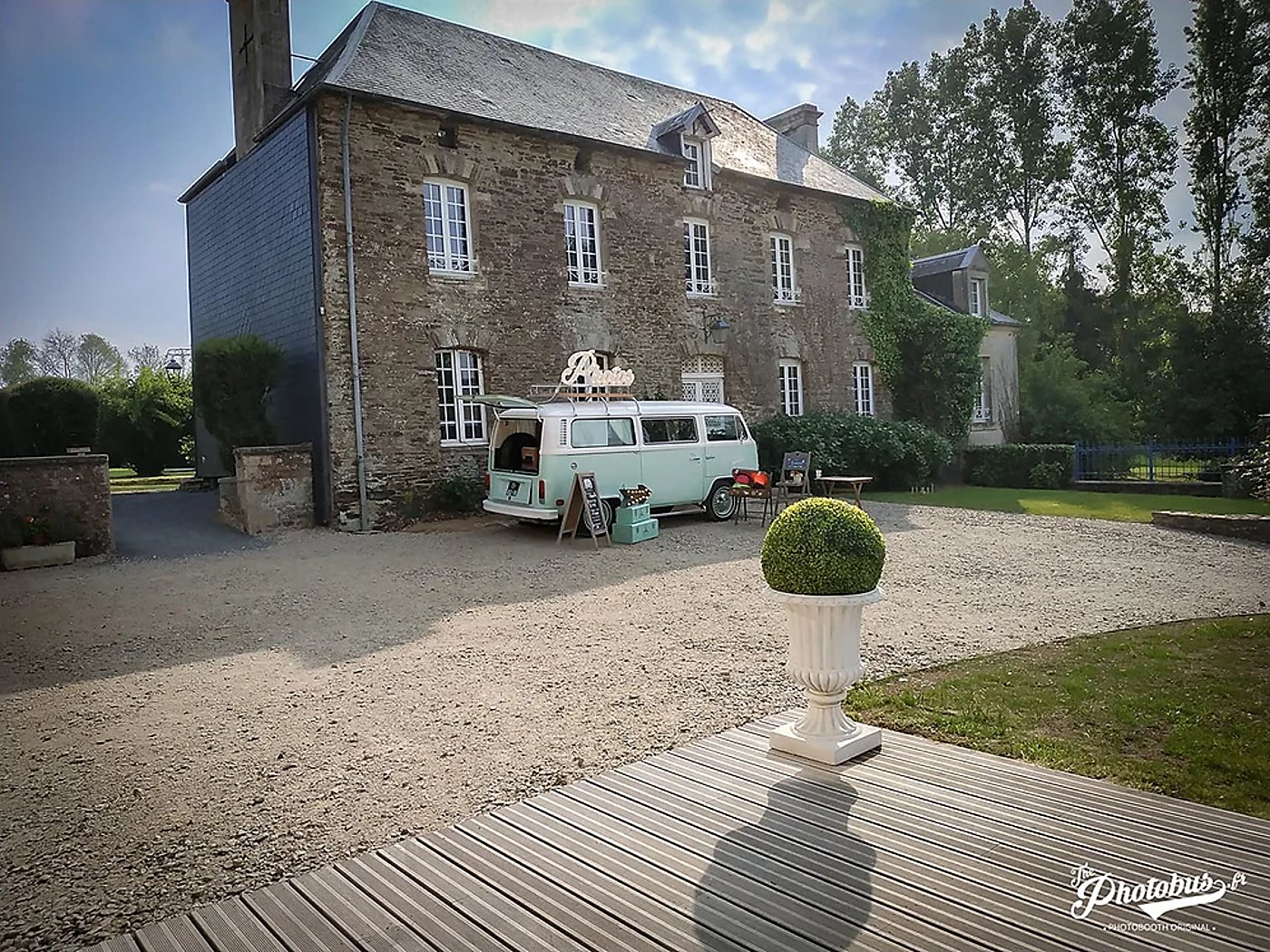 Un combi Volkswagen vert pastel aménagé en photobooth est garé sur le gravier devant un manoir en pierre, avec un buis dans une vasque blanche au premier plan.