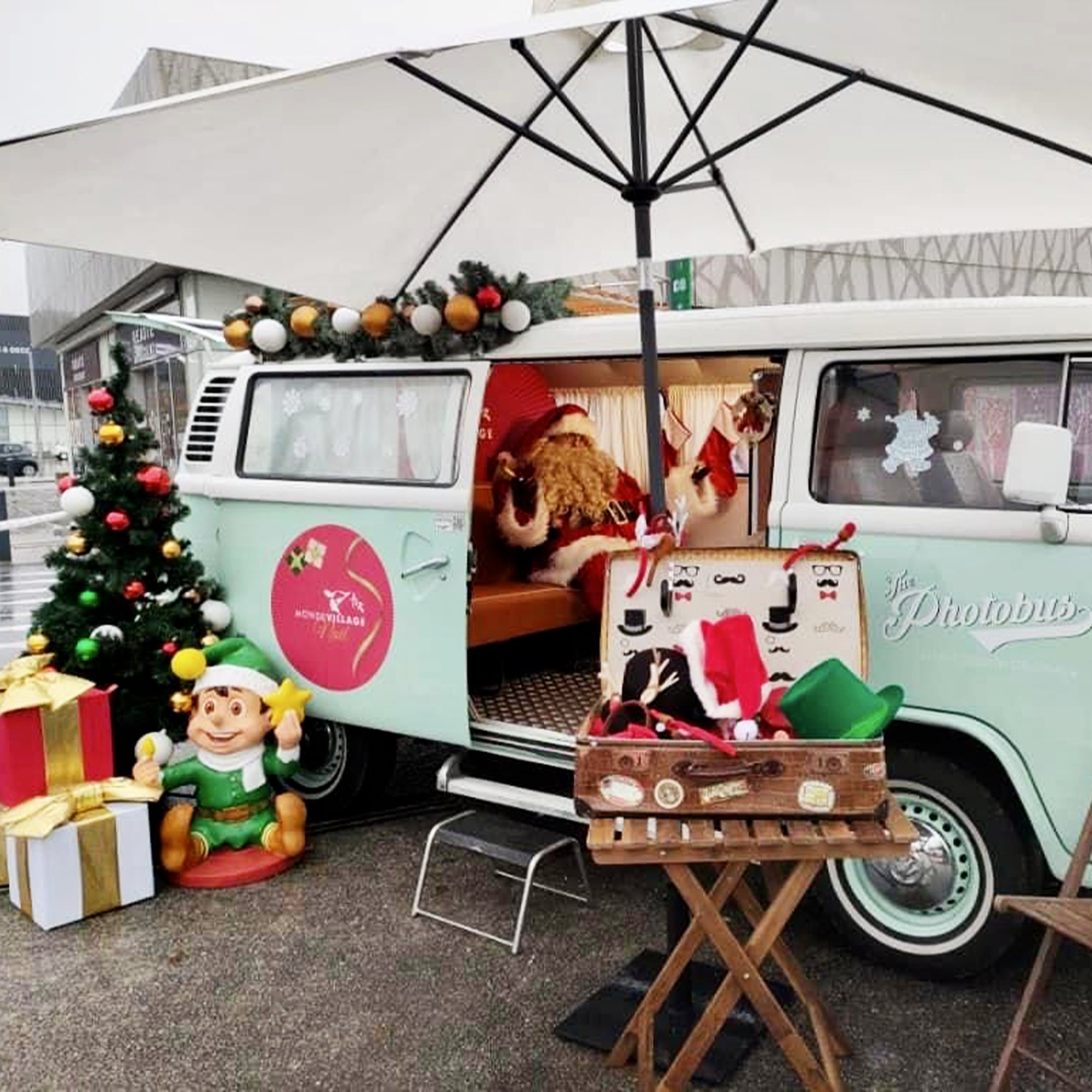 Un combi Volkswagen vert menthe nommé The Photobus, décoré pour Noël, avec un Père Noël assis à l'intérieur, un sapin, des cadeaux, un lutin et une valise d'accessoires photo.