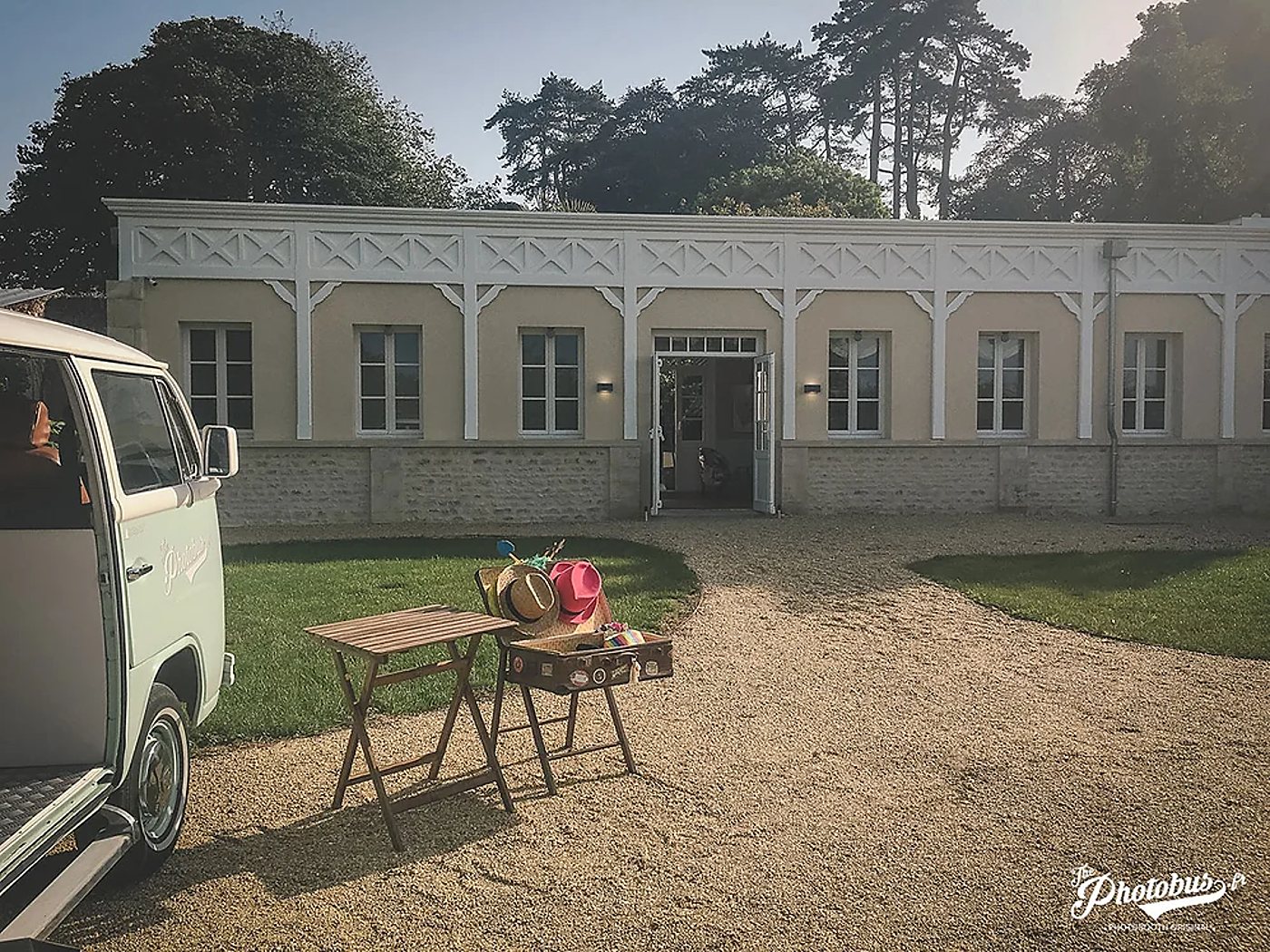 Un combi Volkswagen vintage vert pâle est garé sur le gravier devant un pavillon de plain-pied, avec une table en bois et une valise ouverte remplie de chapeaux colorés.