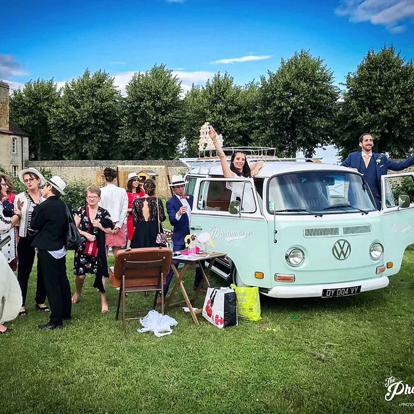 Des invités et les mariés s'amusent autour d'un combi Volkswagen vert menthe aménagé en photobooth sur une pelouse, devant une rangée d'arbres.