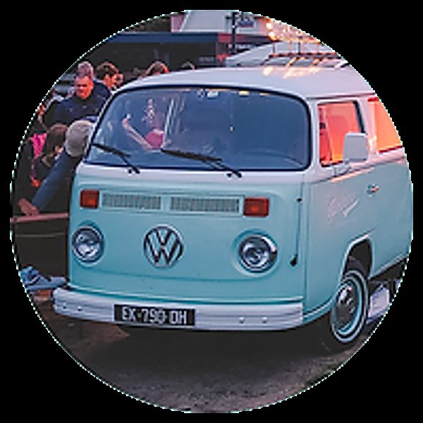 Un combi Volkswagen ancien de couleur bleu pastel et blanc, vu de face, avec des personnes assises en arrière-plan.