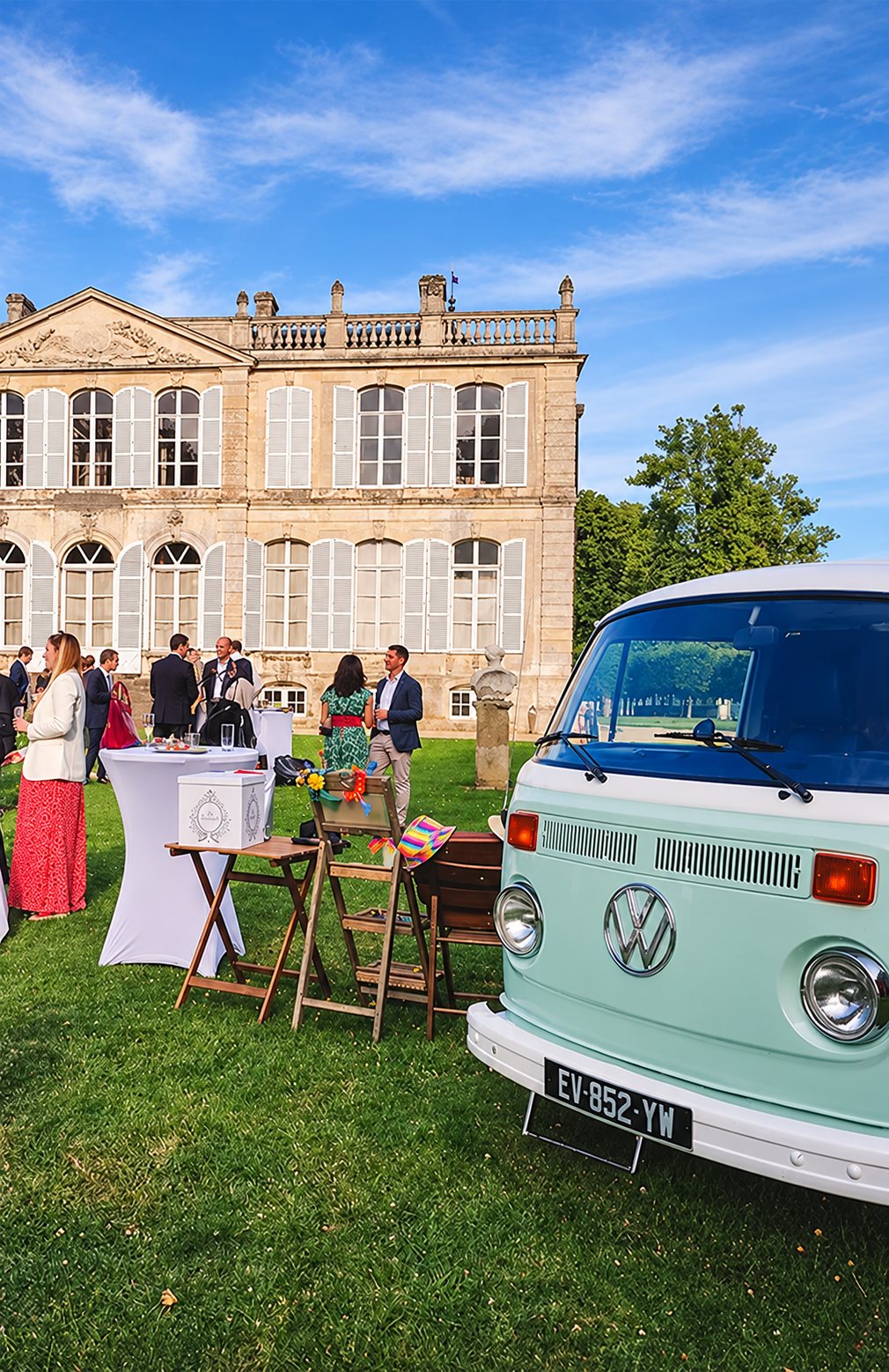 Un combi Volkswagen vert pastel est garé sur la pelouse devant un château, où des invités se réunissent autour de tables hautes lors d'une réception.