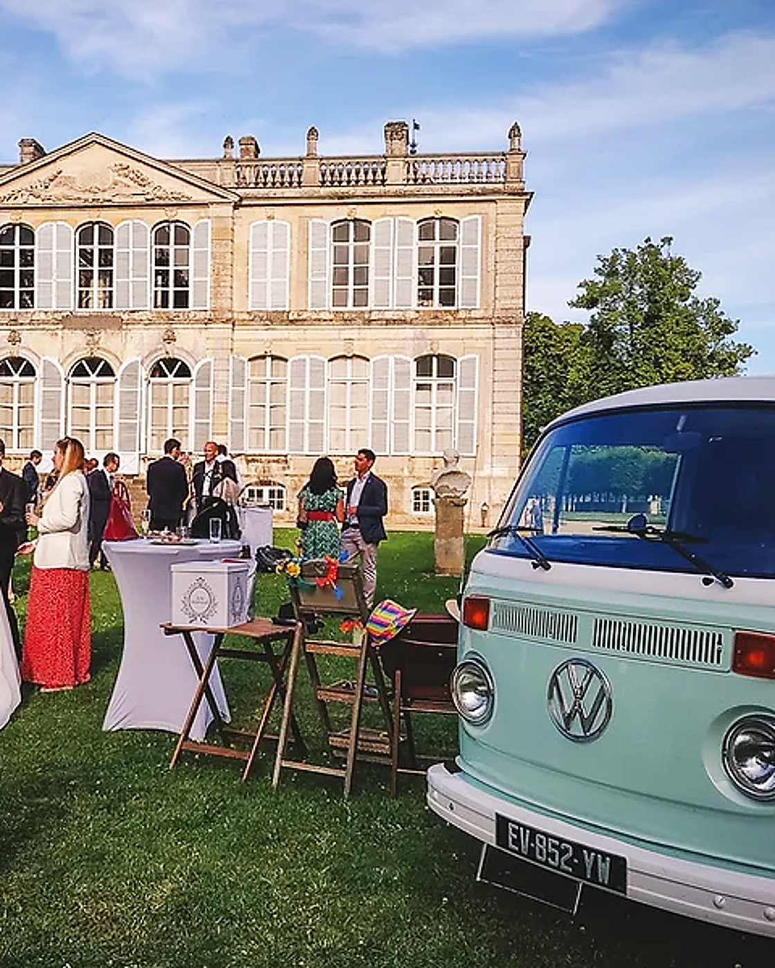 Un combi Volkswagen vert pastel est garé sur la pelouse devant un château où des invités discutent lors d'une réception.