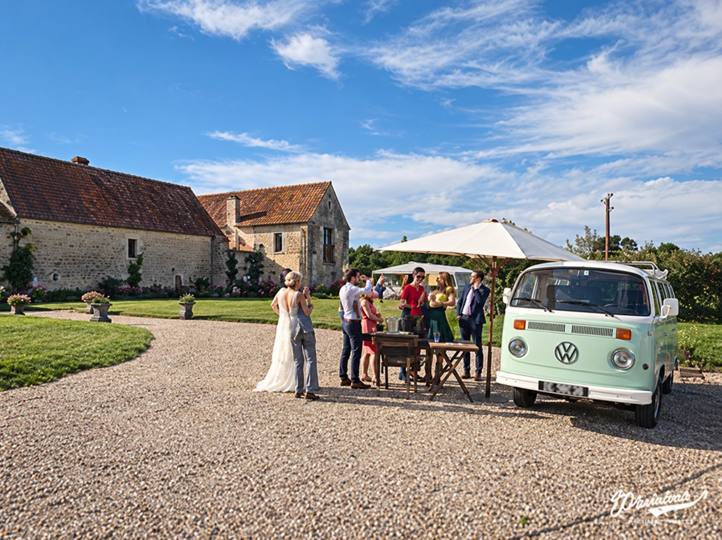 Une mariée en robe blanche et des invités se tiennent près d'un combi Volkswagen vintage vert et blanc sur une cour de gravier, devant une ancienne demeure de pierre.