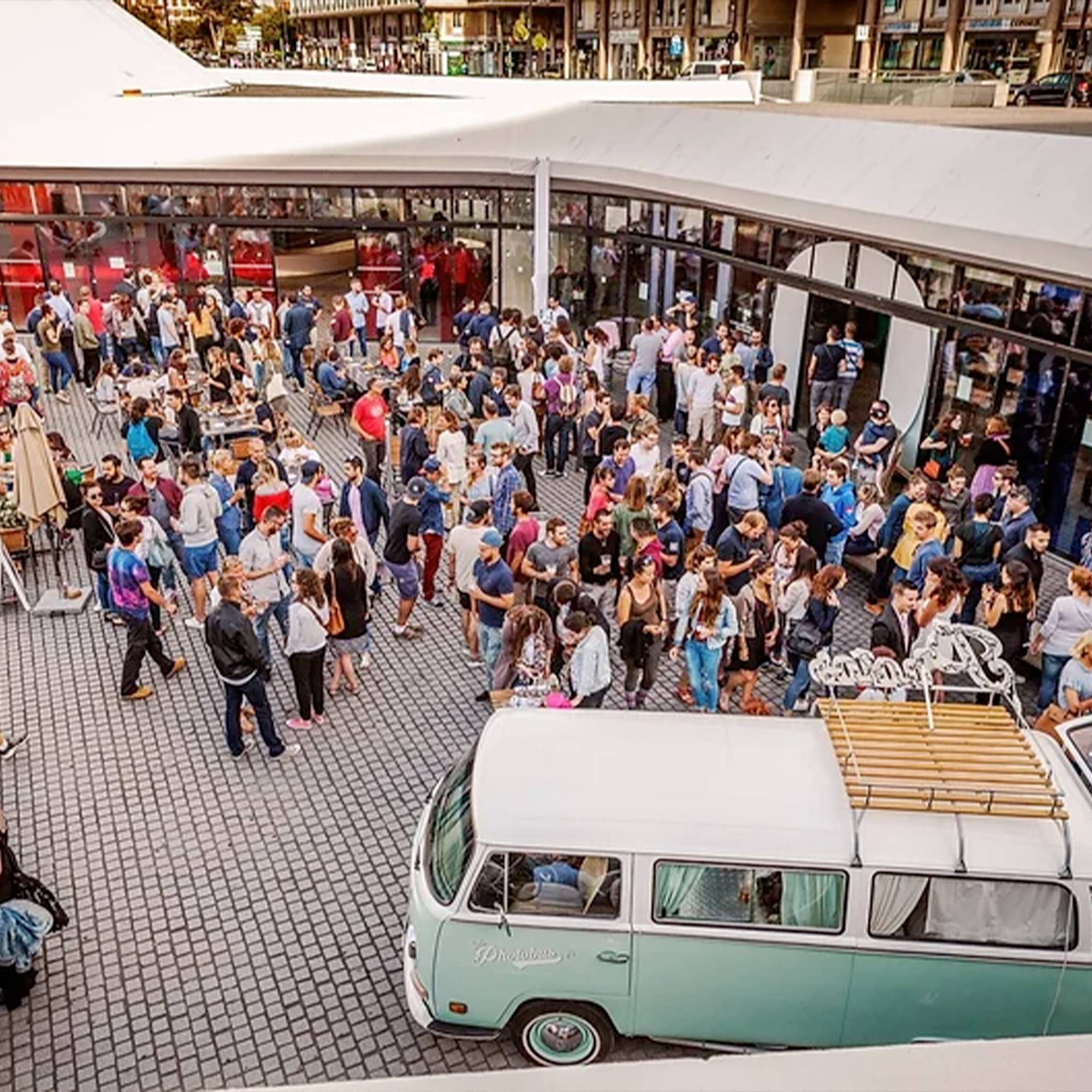 Vue en hauteur d'une foule nombreuse rassemblée sur une place pavée devant un bâtiment vitré, avec un combi Volkswagen vert d'eau et blanc au premier plan.