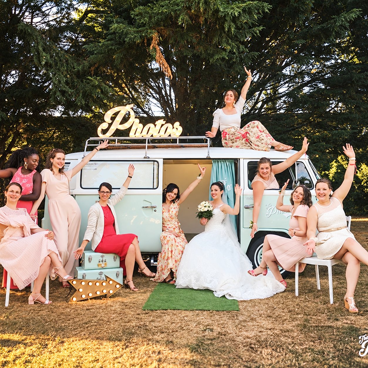 Une mariée entourée de ses demoiselles d'honneur lève les bras devant un combi Volkswagen vintage transformé en photobooth, surmonté d'une enseigne lumineuse « Photos ».