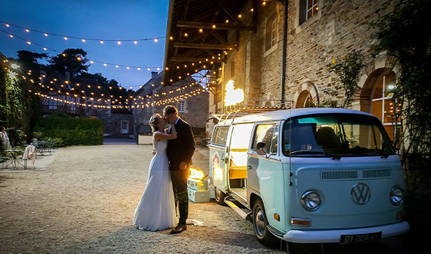 Un couple de mariés s'embrasse dans une cour en gravier, à côté d'un combi Volkswagen bleu clair, sous des guirlandes lumineuses à la tombée de la nuit.