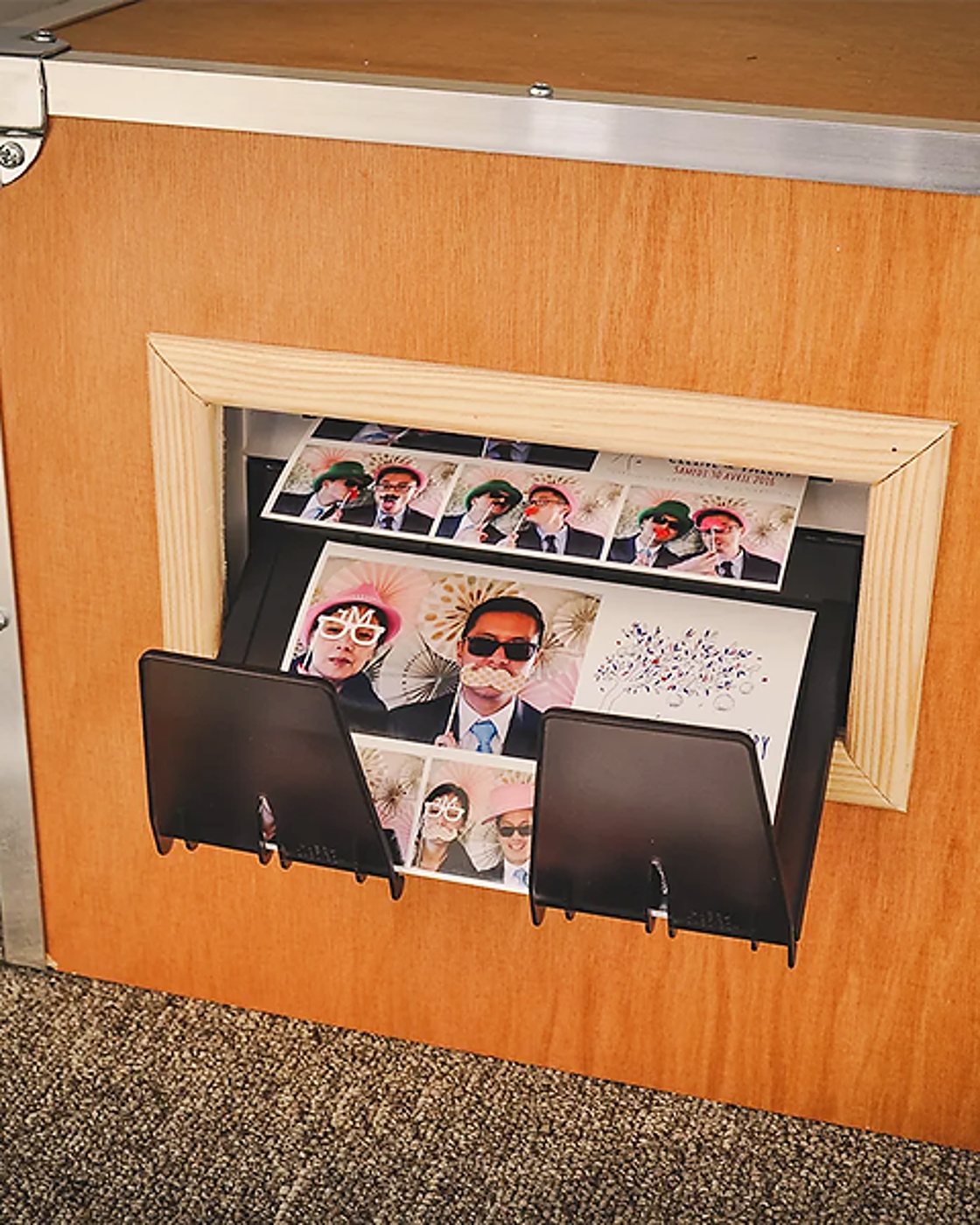 Des tirages de photomaton montrant des invités avec lunettes et accessoires fantaisie sortent d'une fente encadrée de bois sur une borne en bois.